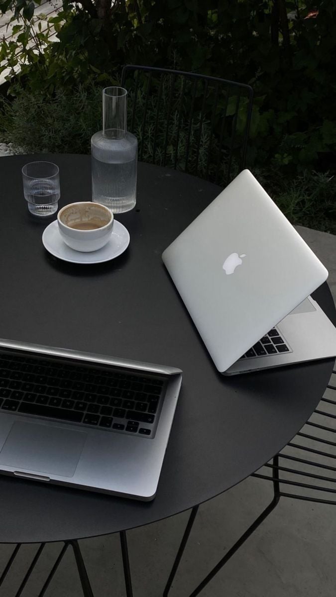 Outdoor workspace with two laptops, coffee cup, and water glass on a round black table surrounded by lush green foliage